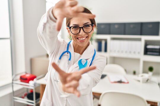 Young brunette doctor woman wearing stethoscope at the clinic smiling making frame with hands and fingers with happy face. creativity and photography concept.