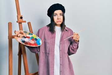 Young brunette woman standing by empty easel stand holding palette depressed and worry for...