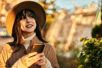 Brunette woman wearing winter hat smiling using smartphone at the park