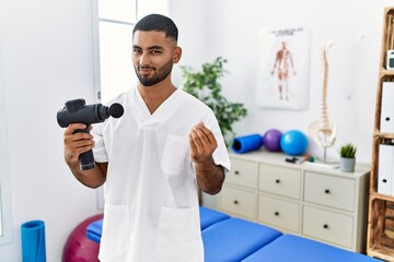 Young indian physiotherapist holding therapy massage gun at wellness center doing money gesture...