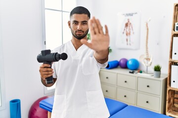 Young indian physiotherapist holding therapy massage gun at wellness center doing stop sing with...