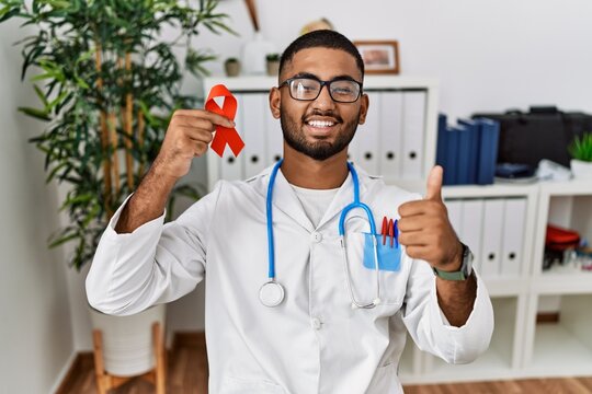 Young indian doctor holding support red ribbon smiling happy and positive, thumb up doing excellent and approval sign