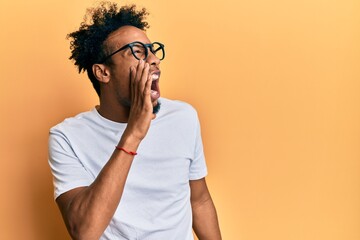 Young african american man with beard wearing casual white t shirt and glasses shouting and screaming loud to side with hand on mouth. communication concept.