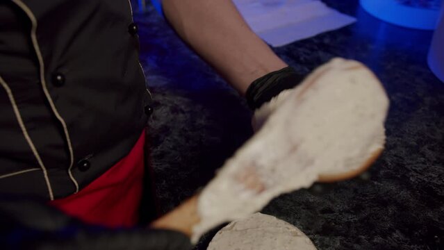 The hands of a chef in black gloves spread white sauce on half a bun for a burger with a wooden spatula.
