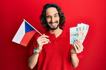 Young hispanic man holding czech republic flag and koruna banknotes winking looking at the camera...