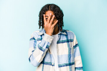 Obraz na płótnie Canvas Young African American man isolated on blue background blink at the camera through fingers, embarrassed covering face.