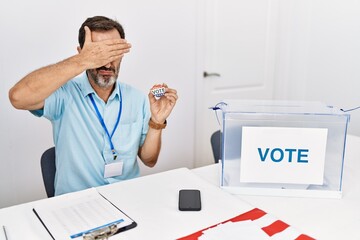 Middle age man with beard sitting by ballot holding i vote badge covering eyes with hand, looking serious and sad. sightless, hiding and rejection concept