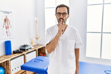 Middle age man with beard working at pain recovery clinic asking to be quiet with finger on lips. silence and secret concept.