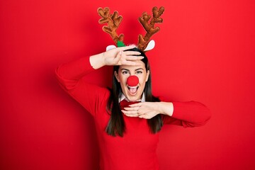 Young hispanic woman wearing deer christmas hat and red nose smiling cheerful playing peek a boo...