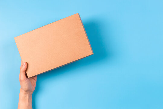 Top view to female hand holding brown cardboard box on light blue background. Mockup parcel box. Packaging, shopping, delivery concept