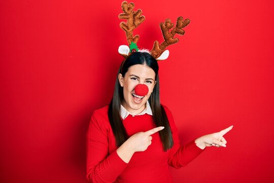 Young hispanic woman wearing deer christmas hat and red nose smiling and looking at the camera pointing with two hands and fingers to the side.