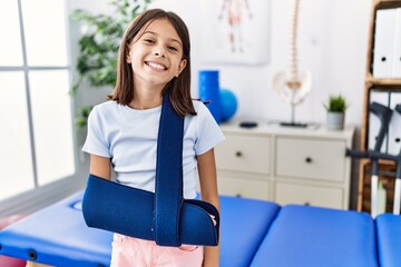 Young hispanic girl wearing arm on sling at rehabilitation clinic looking positive and happy...