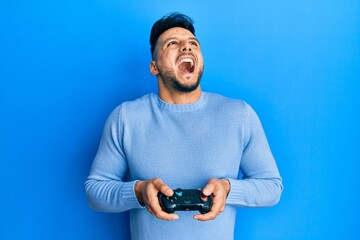 Young arab man playing video game holding controller angry and mad screaming frustrated and furious, shouting with anger looking up.