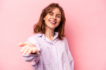 Young caucasian woman isolated on pink background stretching hand at camera in greeting gesture.