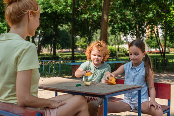 woman sitting near children playing with toys and sand in park.