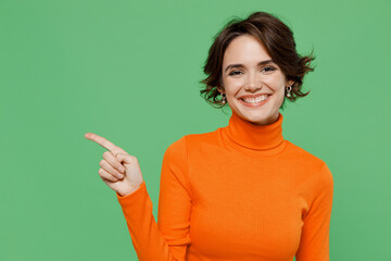 Young smiling woman 20s in casual orange turtleneck point index finger aside on workspace area mock...