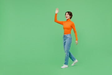 Full body young smiling happy cool fun woman 20s wear casual orange turtleneck walk going waving hand isolated on plain pastel light green color background studio portrait. People lifestyle concept.