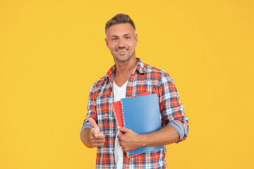 Happy man in casual pointing finger ahead holding school books yellow background, student