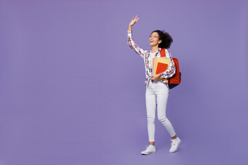 Full body smiling fun young girl woman of African American ethnicity teen student in shirt walk go waving hand hold books isolated on plain purple background. Education in university college concept.