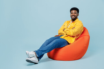 Full body smiling happy young man of African American ethnicity in yellow shirt sit in bag chair...