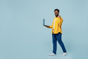 Full body young man of African American ethnicity 20s wear yellow shirt hold use work on laptop pc computer do winner gesture isolated on plain pastel light blue background. People lifestyle concept