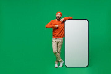 Full body young smiling fun man 20s wear orange sweatshirt hat point index finger on big blank screen mobile cell phone with workspace copy space mockup area isolated on plain green background studio.