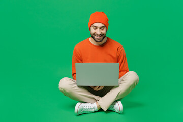Full body young smiling happy man 20s wear orange sweatshirt hat sit hold use work on laptop pc...