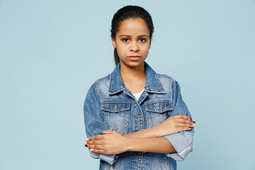 Little serious calm kid teen girl of African American ethnicity 12-13 years old in denim jacket hold hands crossed folded look camera isolated on pastel plain light blue background. Childhood concept.