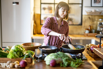 Young woman frying vegetables on cooking pan while cooking healthy food in the kitchen at home. Lots of fresh food ingredients on table. Modern kitchen interior - obrazy, fototapety, plakaty