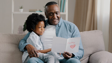 Mature african american man with cute kid girl hugging sitting in room on sofa father reads...