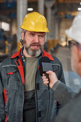 Mature male worker in hardhat and uniform talking in microphone while giving interview about machine factory to reporter