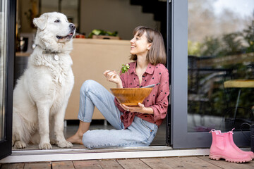Happy woman eats salad while sitting with her adorable huge white dog on terrace near the window. Concept of friendship with pets and healthy eating - Powered by Adobe