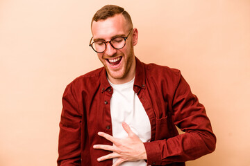 Young caucasian man isolated on beige background laughs out loudly keeping hand on chest.