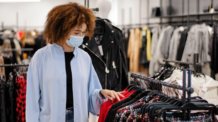 Young african woman wearing safety medical mask choosing clothes in rack in cloth shop. Multiracial curly biracial female customer buyer in protective face respirator shopping clothes in retail store