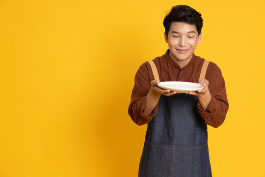 Young Asian man in apron standing and holding empty white plate or dish isolated on yellow background