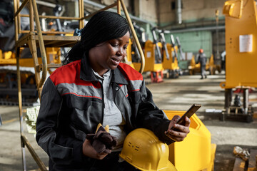 African female worker in uniform reading a message on mobile phone while sitting in warehouse...