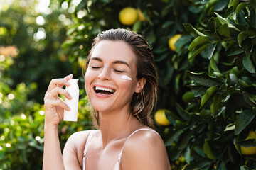 Joyful and beautiful woman applying moisturizing cream or sunblock on her facial skin
