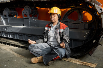 African female worker in uniform sitting in lotus position and meditating with tractor in the...