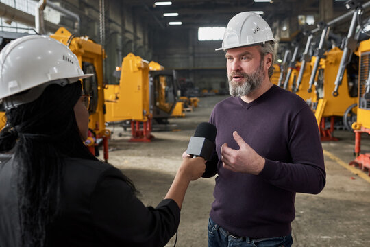 Mature bearded engineer in work helmet speaking in microphone and giving interview to reporter during reportage in plant