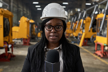 Portrait of African journalist in work helmet speaking fresh news to microphone at plant