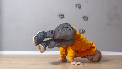 A Caucasian boy in an orange hoodie is standing on his hands with his legs raised up, frozen, the child is dancing a break dance to the room