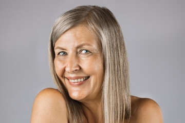 Beautiful aged woman with smooth healthy skin and bare shoulders smiling sincerely on camera while posing over grey studio background. Concept of natural aging.