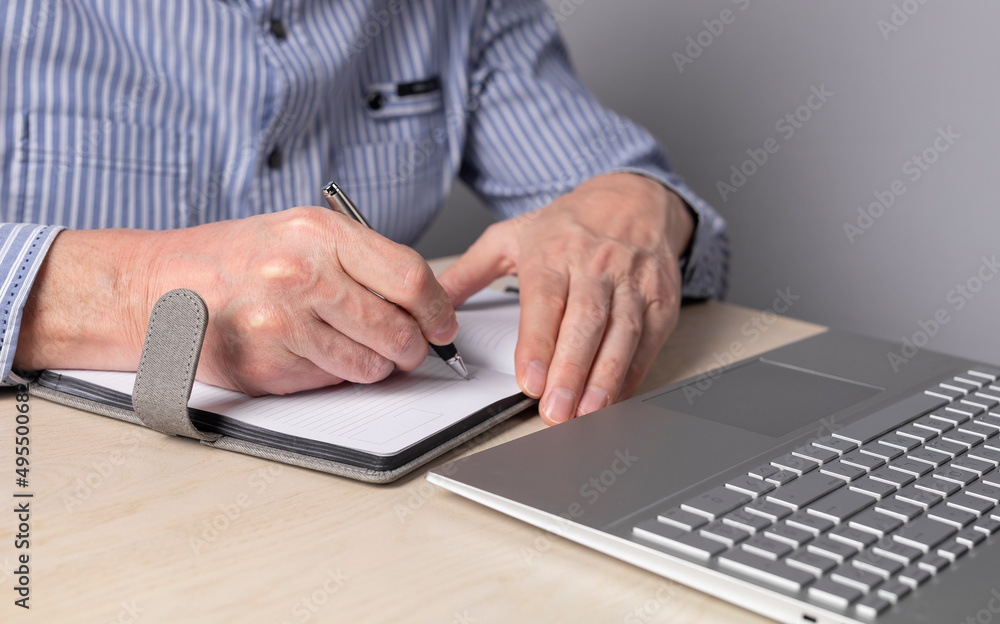 Poster man sitting at table with laptop and writing with pen in notebook. hands closeup. student or freelan