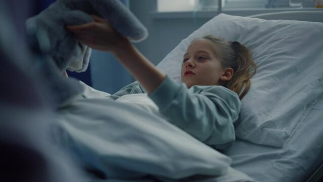 Sick girl playing soft toy in clinic bed. Kid lying in modern hospital room. 