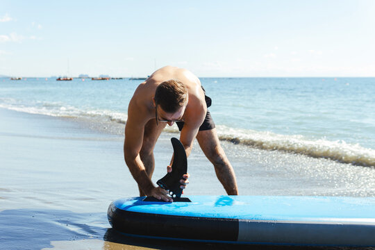 Young male surfer setting up standup paddleboard on a beach.