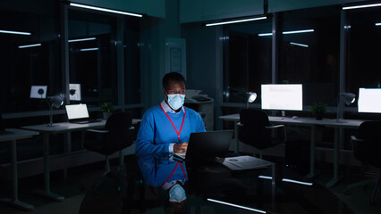 African-American businessman in facemask work on laptop late in dark office