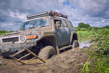 Obraz na płótnie Canvas Off-road vehicle got stuck on impenetrable road after rain during competition. Off-road 4x4 car is stuck in a puddle of mud and is trying to leave using a ladder. The concept of extreme entertainment.