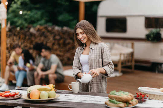 Happy millennial Caucasian woman pouring tea, making breakfast near RV, camping with her diverse friends outdoors