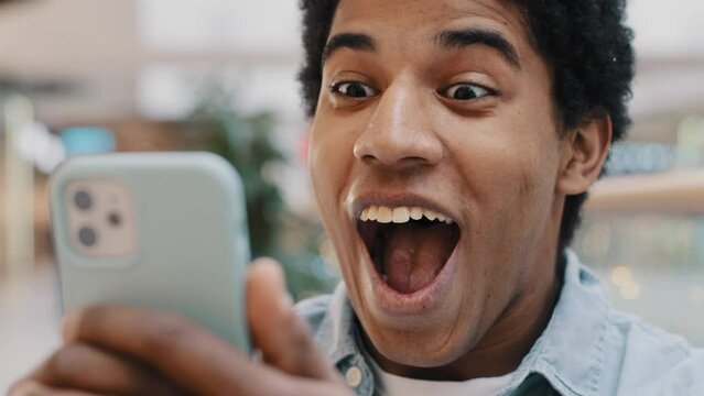African happy excited guy surprised by message good news winning online bets looking at mobile phone has offer invite opens mouth in surprise delighted shock man feels amazing wonder with achievement
