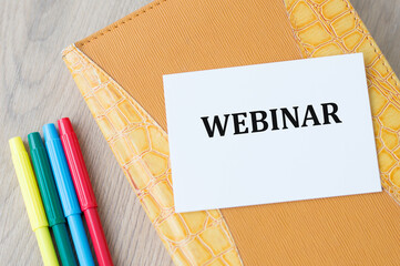 A white business card with Webinar text on a notepad on a wooden table with rows of colored markers